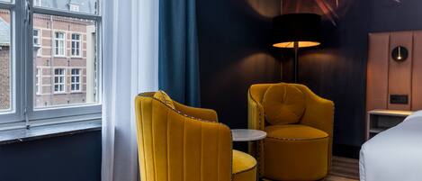 Una habitación de hotel con un ventanal grande, un sillón amarillo, una mesa redonda, una lámpara y una cama.