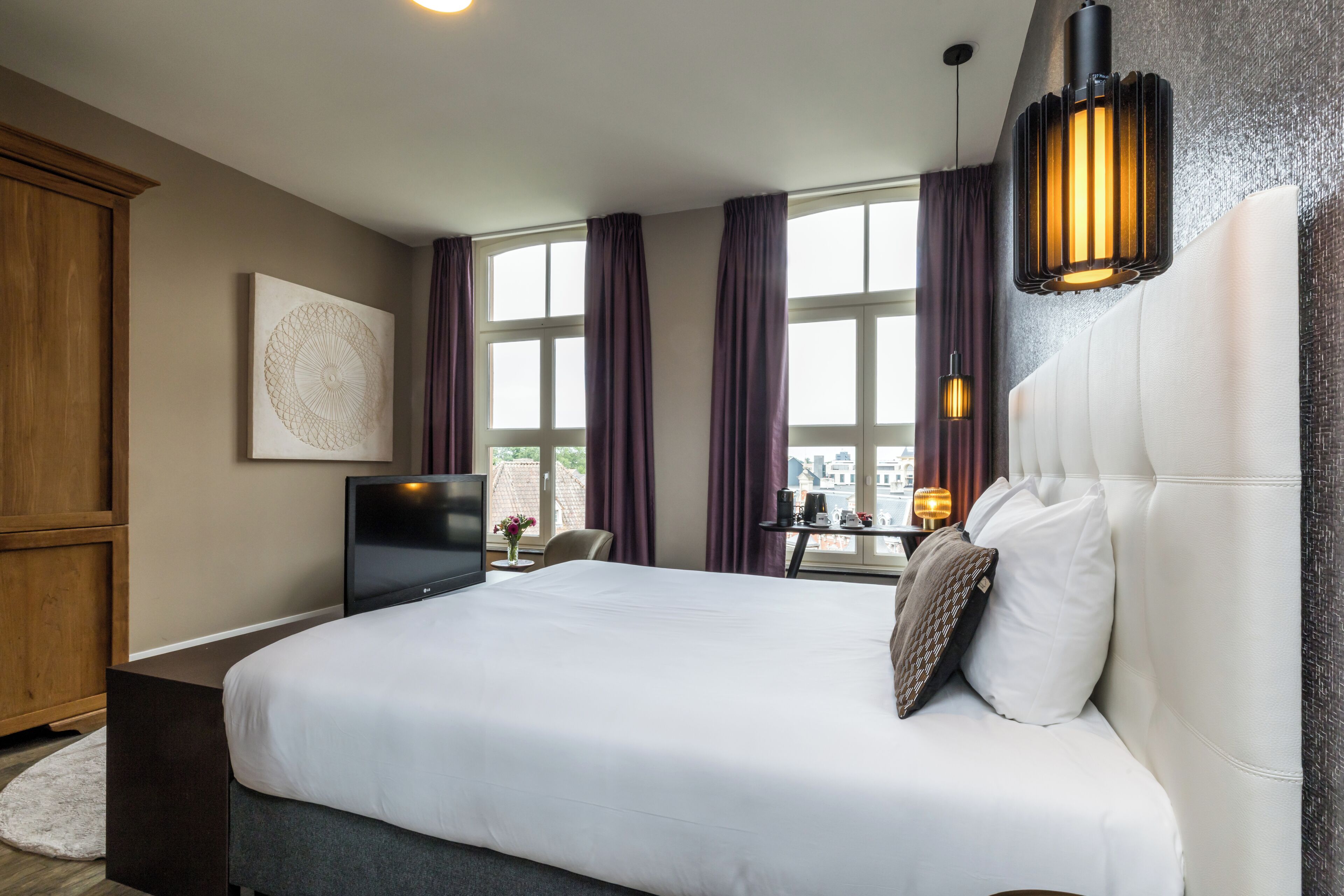 A modern hotel room with a large bed, a flat-screen TV, a dining table, and a view of the city.