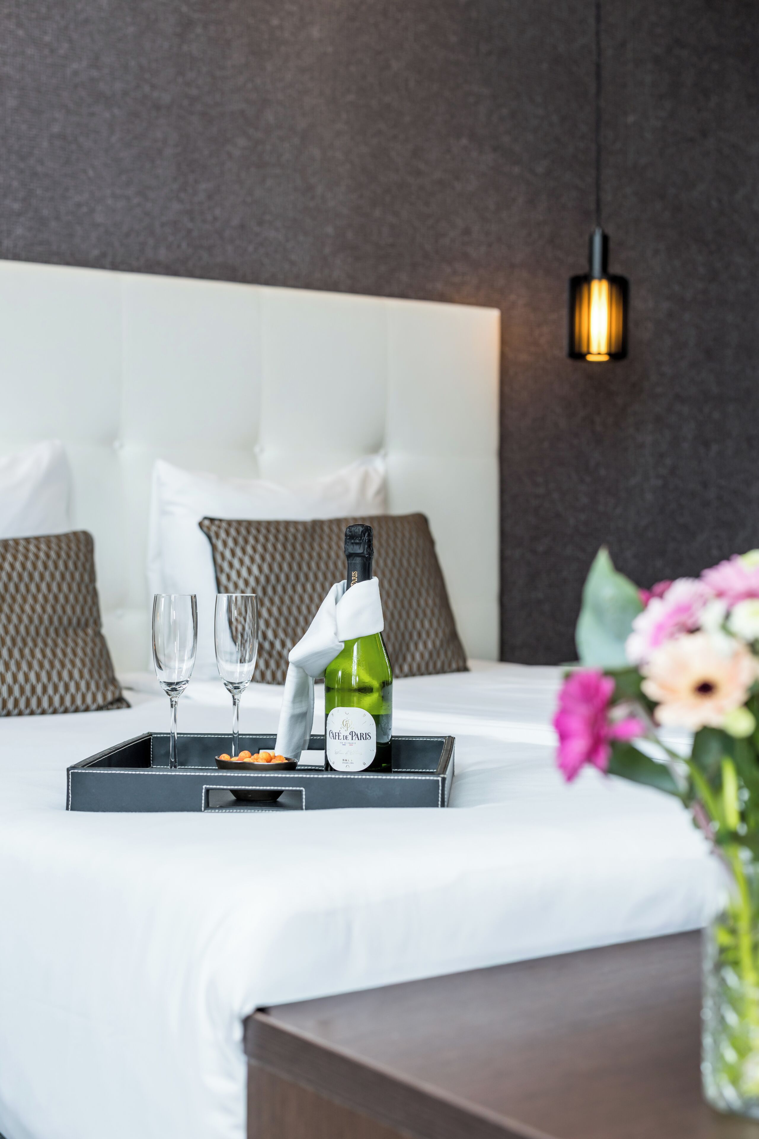 A neatly made bed with a tray containing a bottle of wine, two glasses, and some snacks.