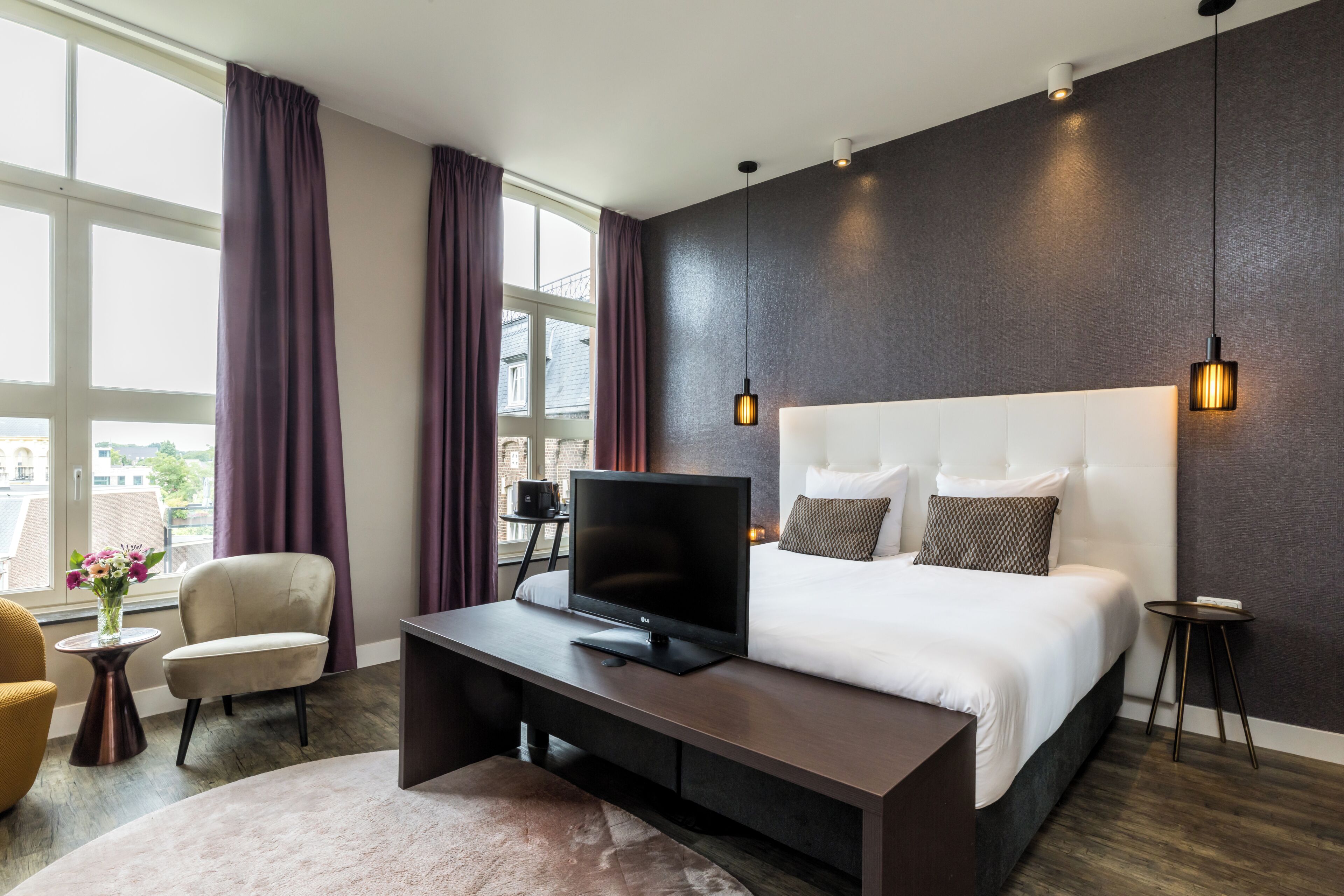 A modern hotel room with a large bed, a flat-screen TV, a desk, and a view of the city through large windows.