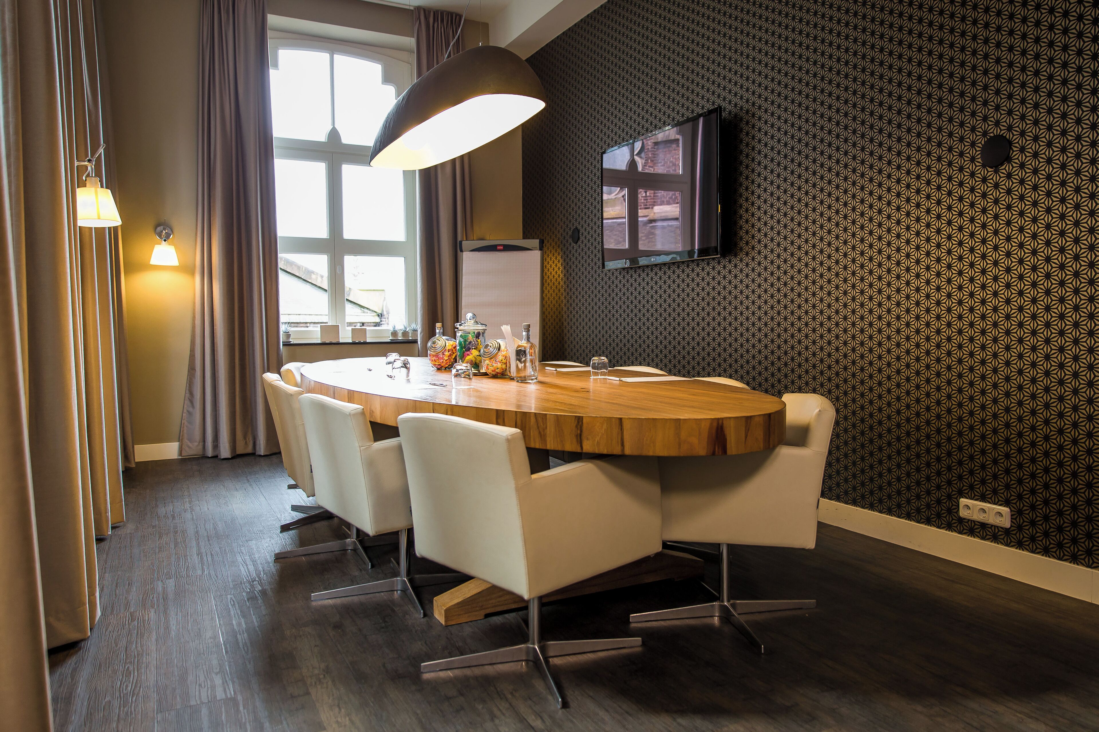A modern dining area with a round wooden table, white chairs, a large pendant light, and a TV mounted on the wall.