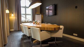 A modern dining area with a round wooden table, white chairs, a large pendant light, and a TV mounted on the wall.