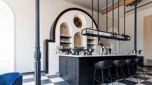 Una zona de bar con piso a cuadros, taburetes negros y estantes con botellas y copas.
