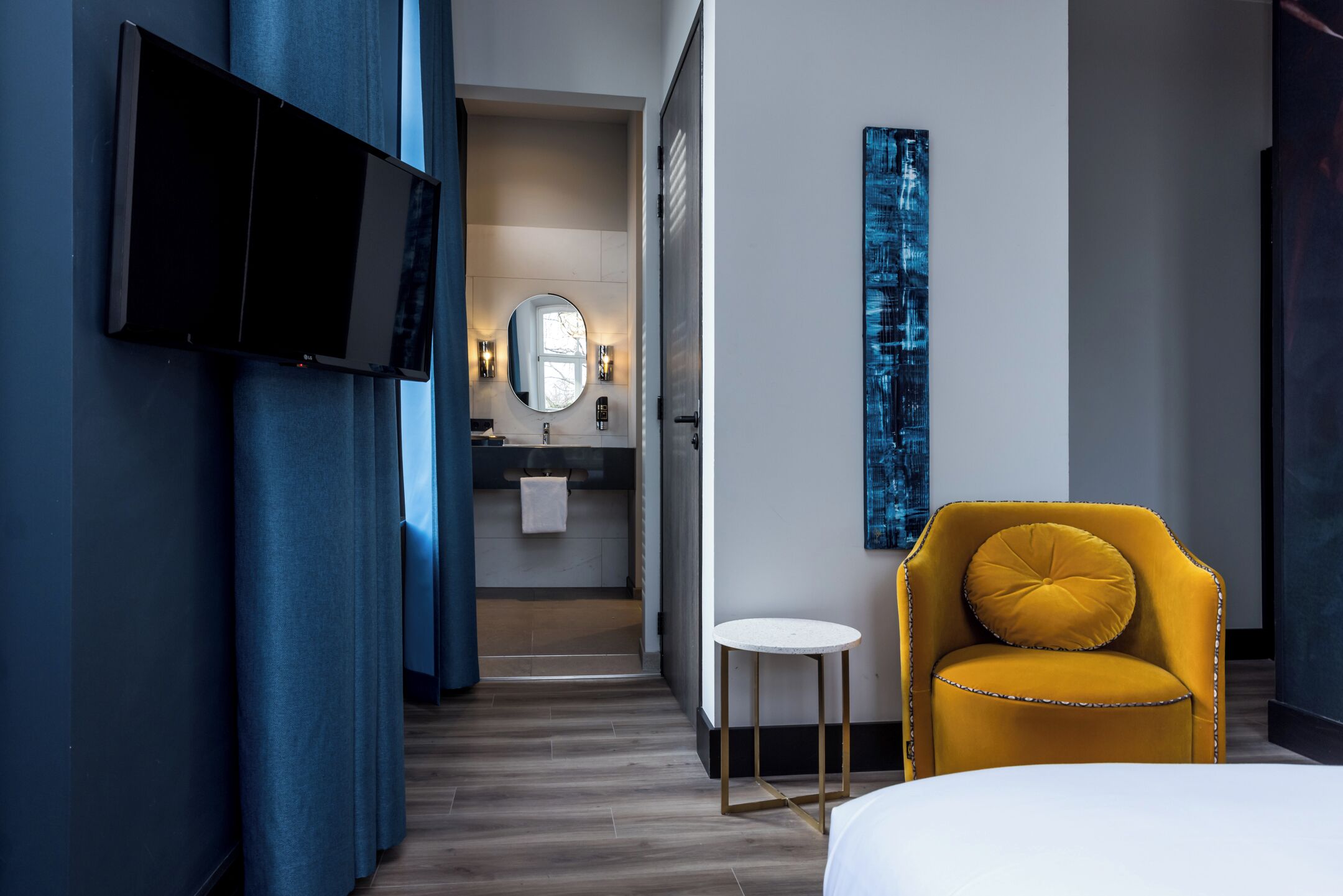 Une chambre d’hôtel moderne avec un grand lit, un téléviseur à écran plat fixé au mur, une petite table ronde et un fauteuil jaune.