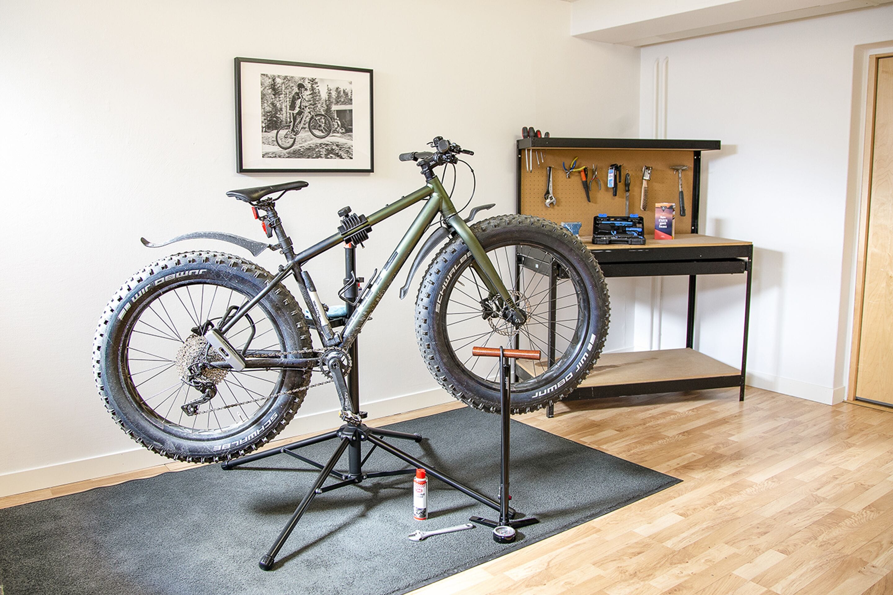 A bicycle mounted on a stand in a room with a wooden floor, a pegboard with tools, and a framed picture on the wall.