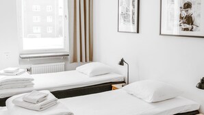 Habitación de hotel con dos camas individuales, ropa de cama blanca, una ventana con cortinas, un radiador y un cuadro en la pared.