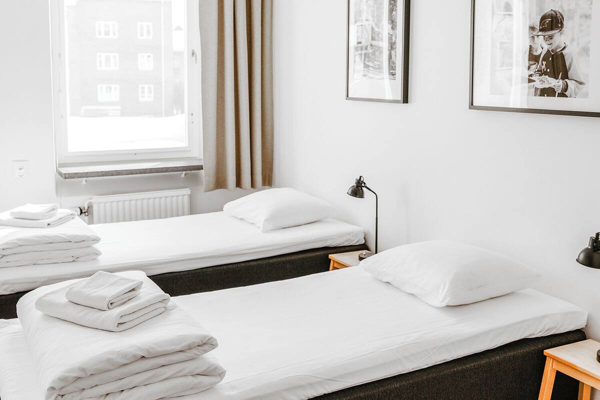 Ein Hotelzimmer mit zwei Einzelbetten, weißen Laken, einem Fenster mit Vorhängen, einer Heizung und einem gerahmten Bild an der Wand.