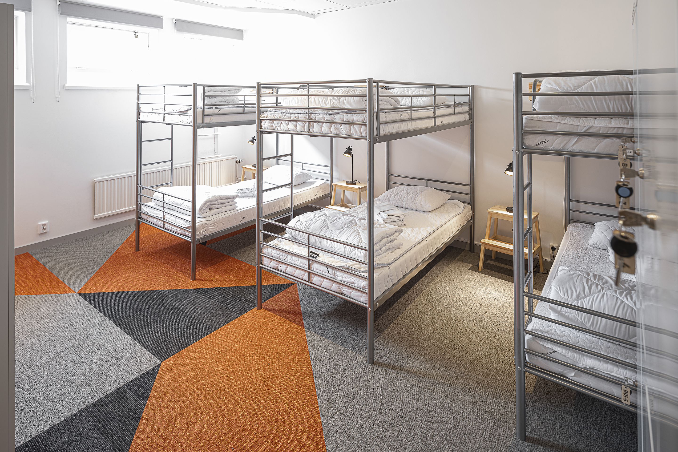 A dormitory room with bunk beds, a small bedside table, a lamp, and a radiator.