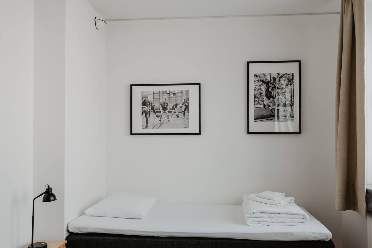 Ein schlicht eingerichteter Raum mit einem Bett, einem kleinen Holztisch mit einer Lampe und zwei gerahmten Bildern an der Wand.
