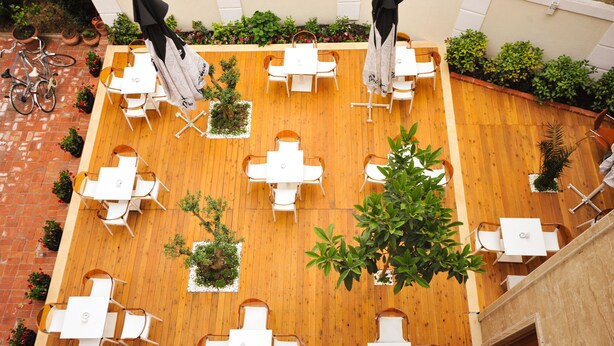 Un patio al aire libre con piso de madera, mesas y sillas blancas, y plantas en macetas.