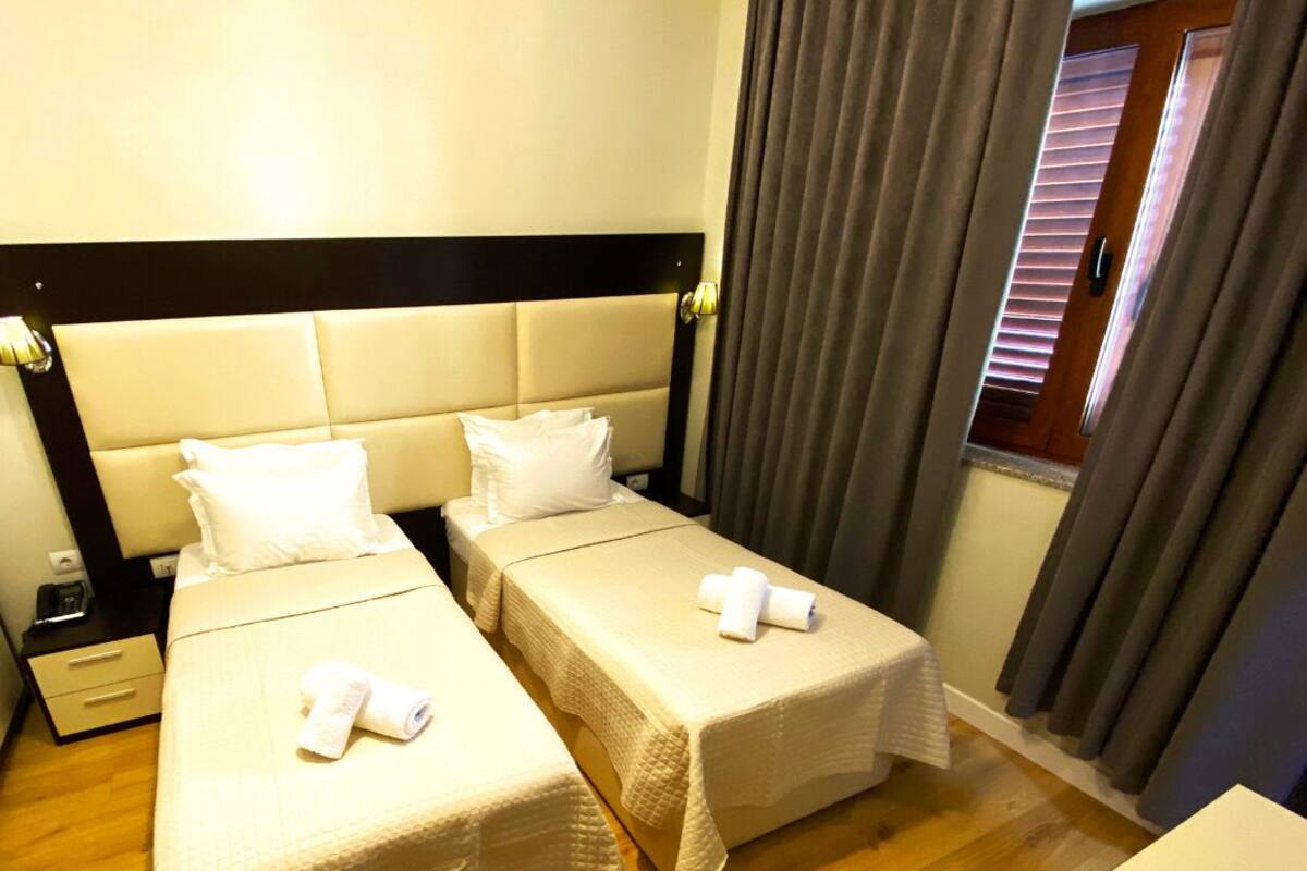 Habitación de hotel con dos camas, cada una con cubrecama y almohadas blancas, suelo de madera y una ventana con cortinas.