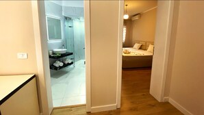 Habitación de hotel con piso de madera, cama, mesita de noche y baño con lavamanos y espejo.