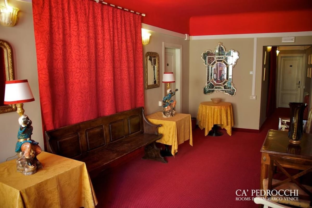 A room with a red carpet, a wooden bench, a small table with a yellow tablecloth, and a mirror on the wall.