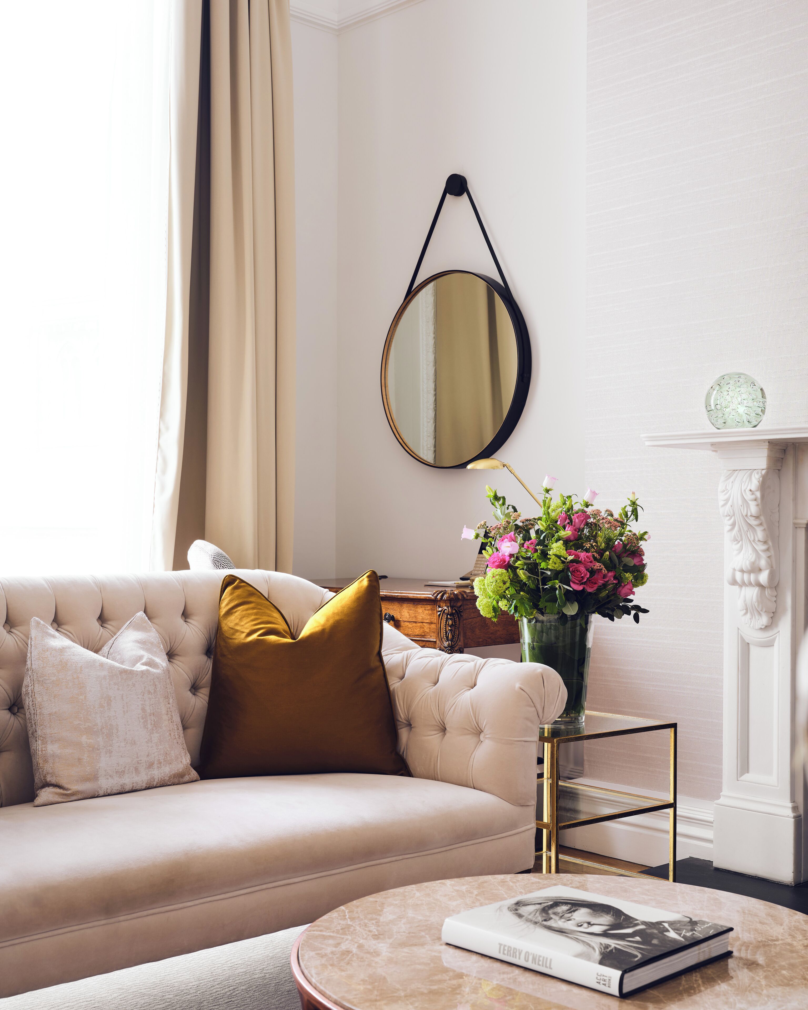 A living room with a beige sofa, a round mirror, a vase with flowers, and a fireplace.