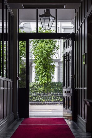 A hallway with a red carpet leading to a door, flanked by dark wooden panels and a hanging light fixture. - The Adria (London)