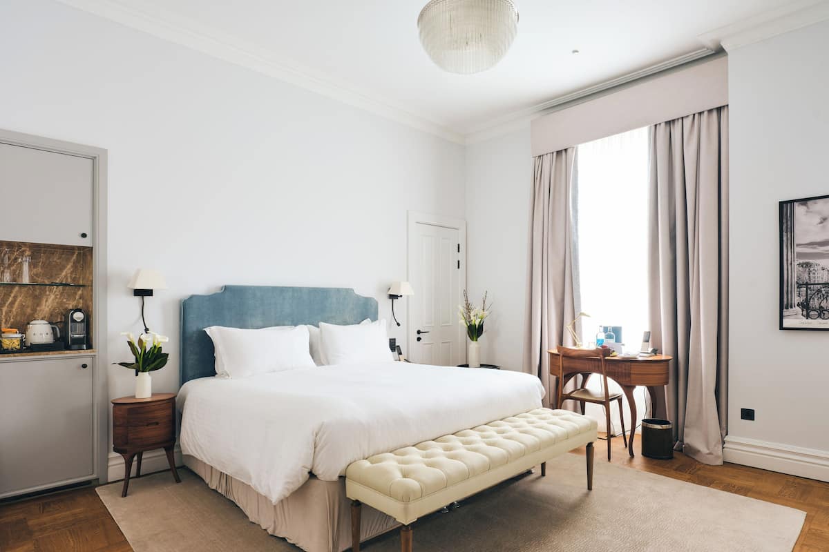 Une chambre à coucher comprenant un lit, des tables de chevet, un banc, un bureau et une fenêtre avec des rideaux.