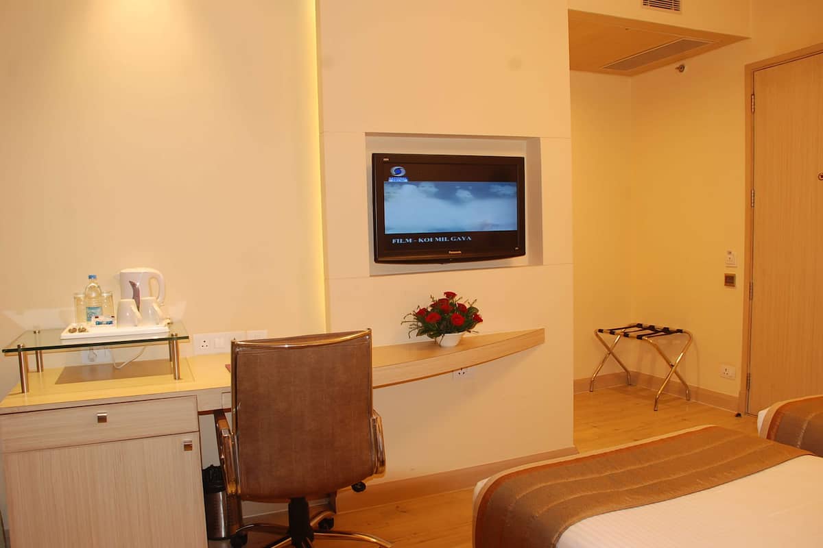 Una habitación de hotel con una cama, un escritorio con una silla, un televisor montado en la pared, una mesita con un florero y un perchero.