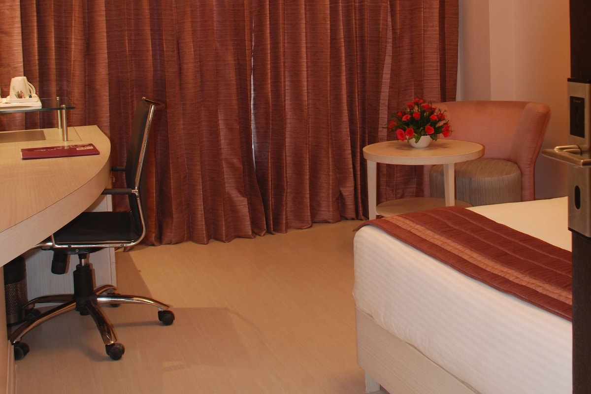 Quarto de hotel com cama, escrivaninha com cadeira, mesa pequena com um vaso de flores e uma janela grande com cortinas.