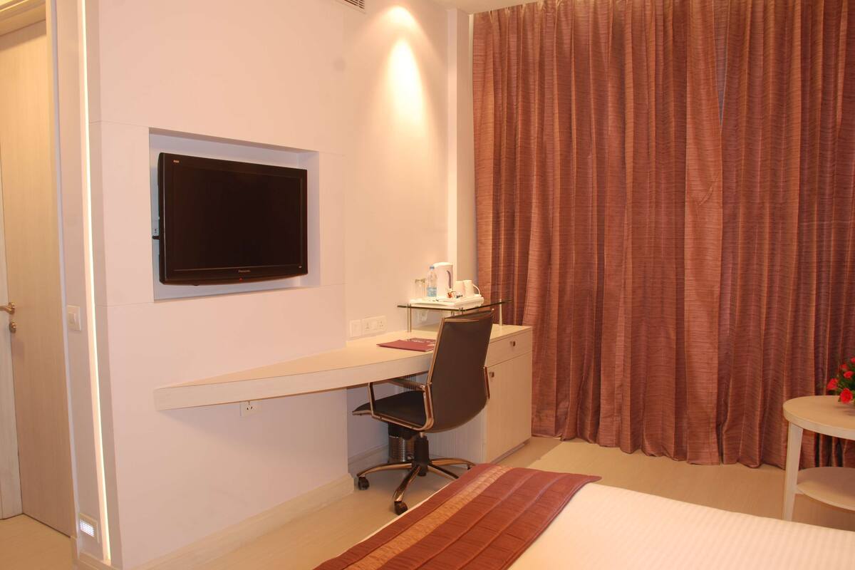 Habitación de hotel con cama, escritorio con silla, televisor montado en la pared y ventana con cortinas.