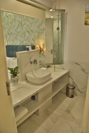 Une salle de bain moderne avec un lavabo en marbre blanc, une cabine de douche en verre et une tête de lit recouverte d’un tissu bleu.