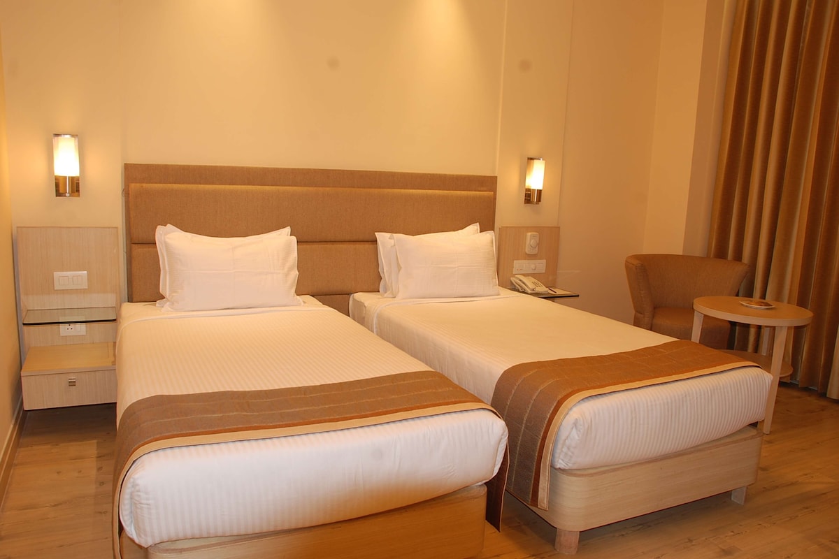 Quarto de hotel com duas camas, uma cadeira, uma mesinha e um abajur.