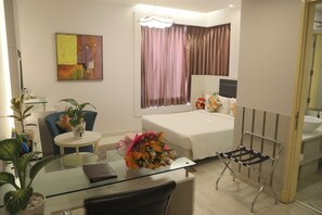 Une chambre d’hôtel avec un lit, un bureau et une chaise, une petite table avec une plante, une fenêtre avec des rideaux et une salle de bain visible à travers une porte ouverte.