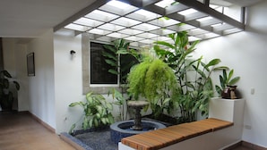 A room with a tiled floor, a white bench with a striped cushion, a small fountain, and various green plants. - Hotel El Almendro (Managua)