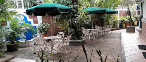 Un patio al aire libre con mesas y sillas de hierro blanco forjado, sombrillas verdes y plantas en macetas.