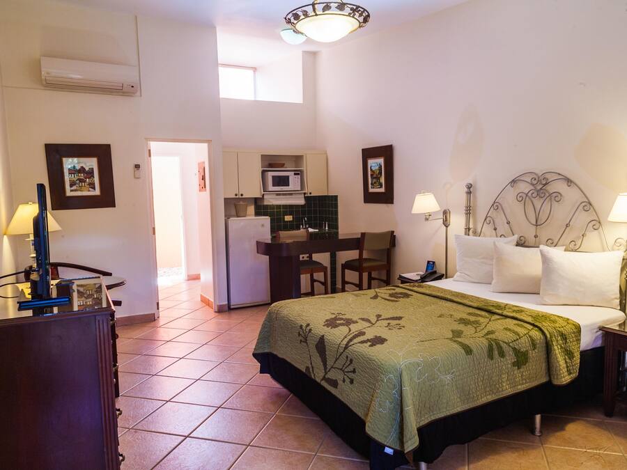 Habitación de hotel compacta con cama, mesita de noche, lámpara y zona de kitchenette.