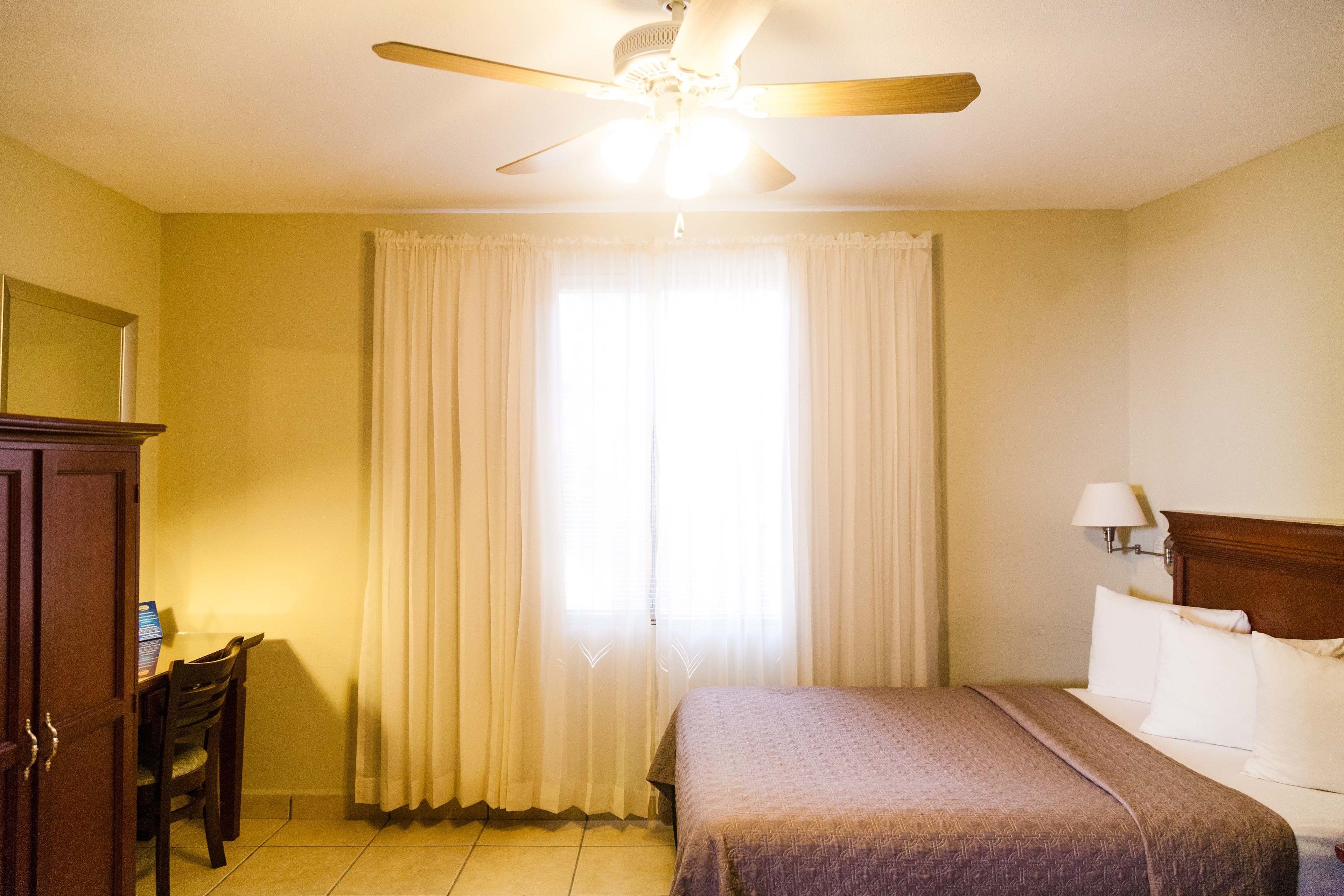 A hotel room with a bed, a desk, a chair, a ceiling fan, and a window with curtains.