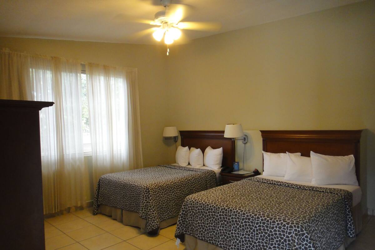 A hotel room with two beds, a ceiling fan, a nightstand with a lamp, and a window with curtains.