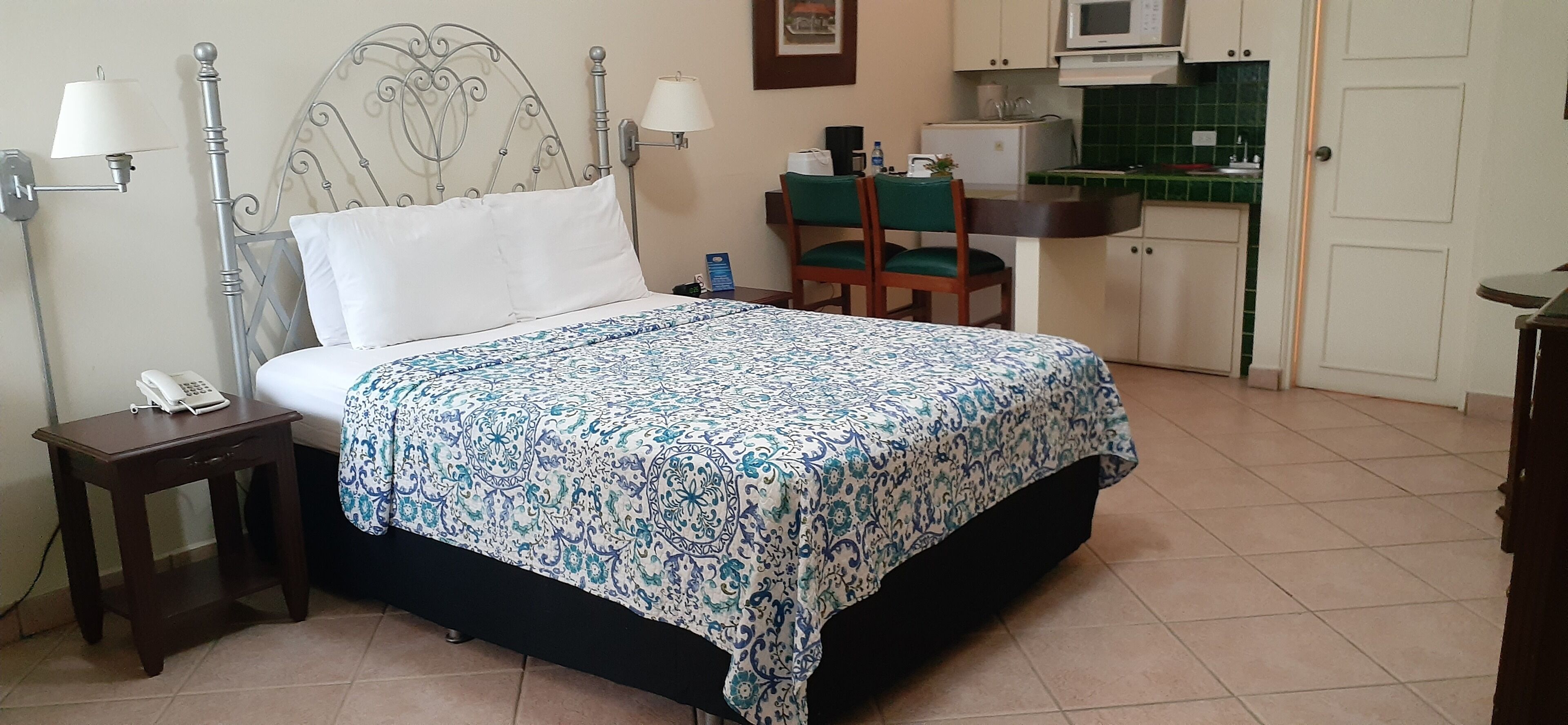 Un dormitorio ordenado con cama, lámparas de noche, teléfono y una pequeña mesa. Junto al dormitorio, hay una kitchenette compacta con microondas, un lavabo y una silla.