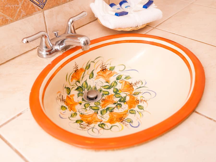 A bathroom sink with an orange rim and floral design, a chrome faucet, and a white towel basket on the floor.