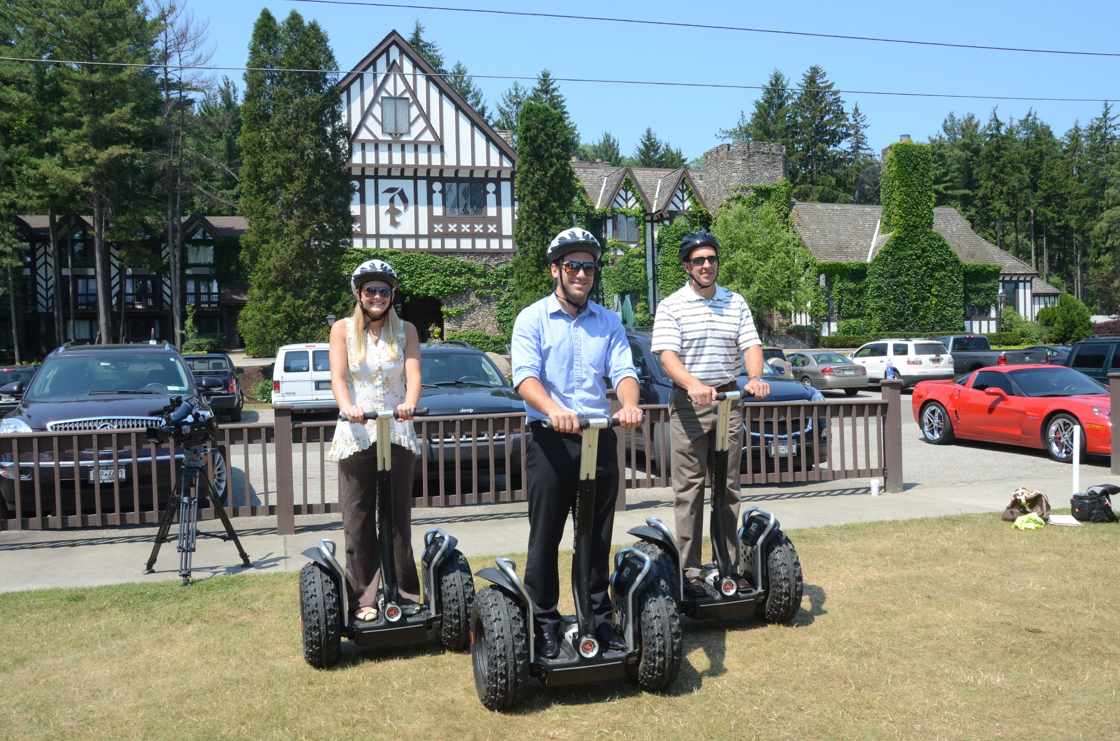 Drei Personen fahren auf Segways über eine Wiese. Im Hintergrund sind parkende Autos und ein Gebäude mit markanter Architektur zu sehen.