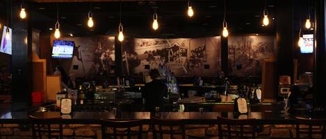 A bar with wooden chairs, hanging light bulbs, and a television screen.