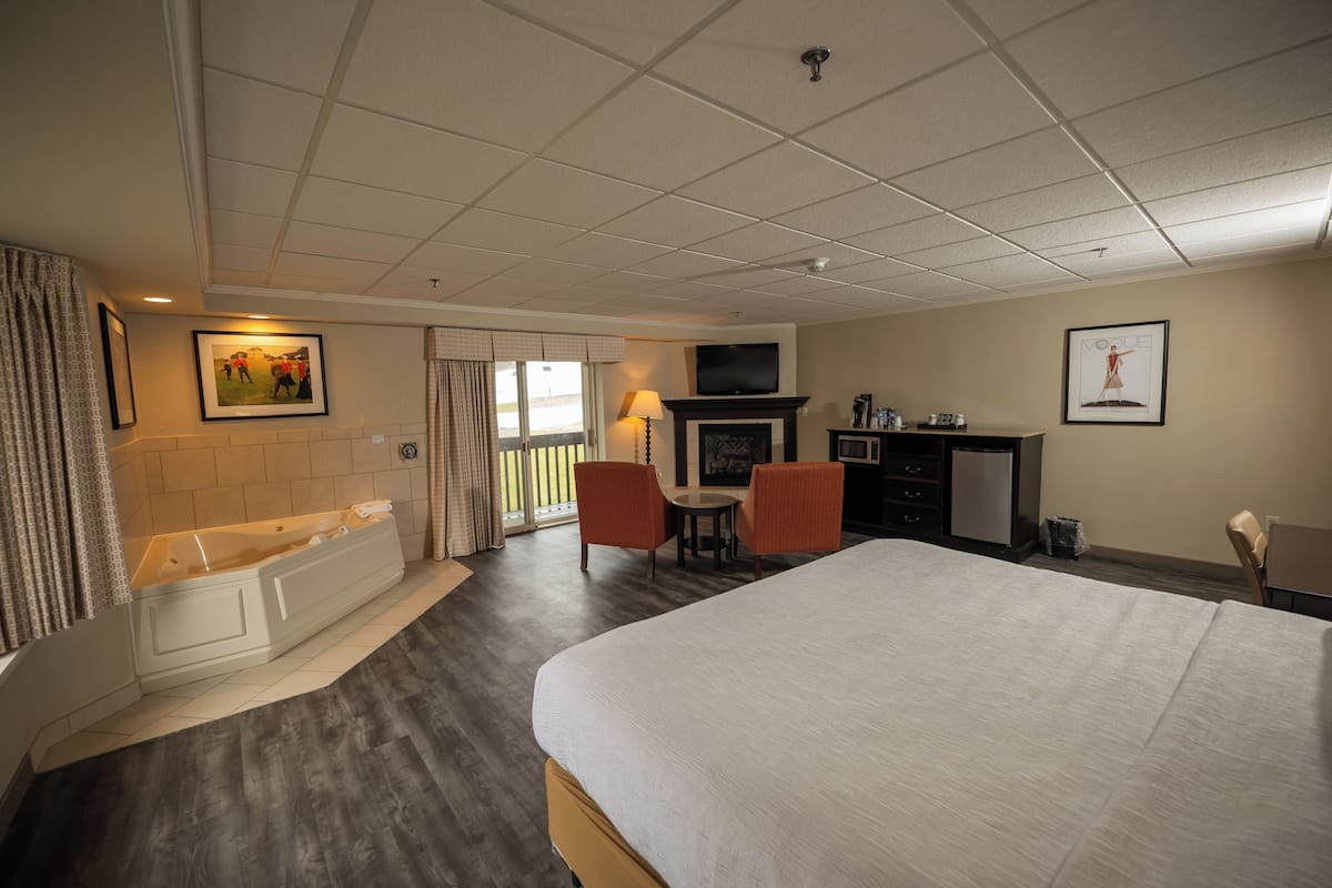 A hotel room with a large bed, a sitting area with a table and chairs, a fireplace, a television, and a balcony with curtains.