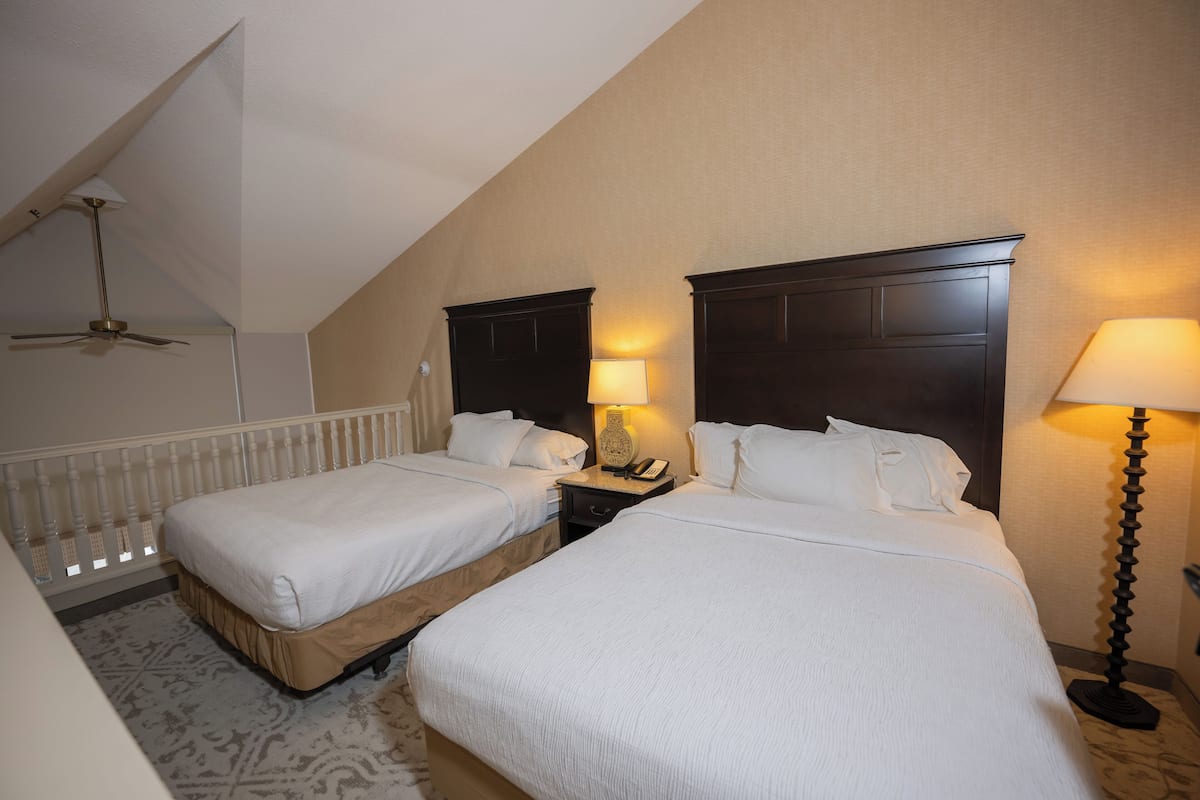 A hotel room with two beds, a ceiling fan, a nightstand with a lamp, and a patterned rug.