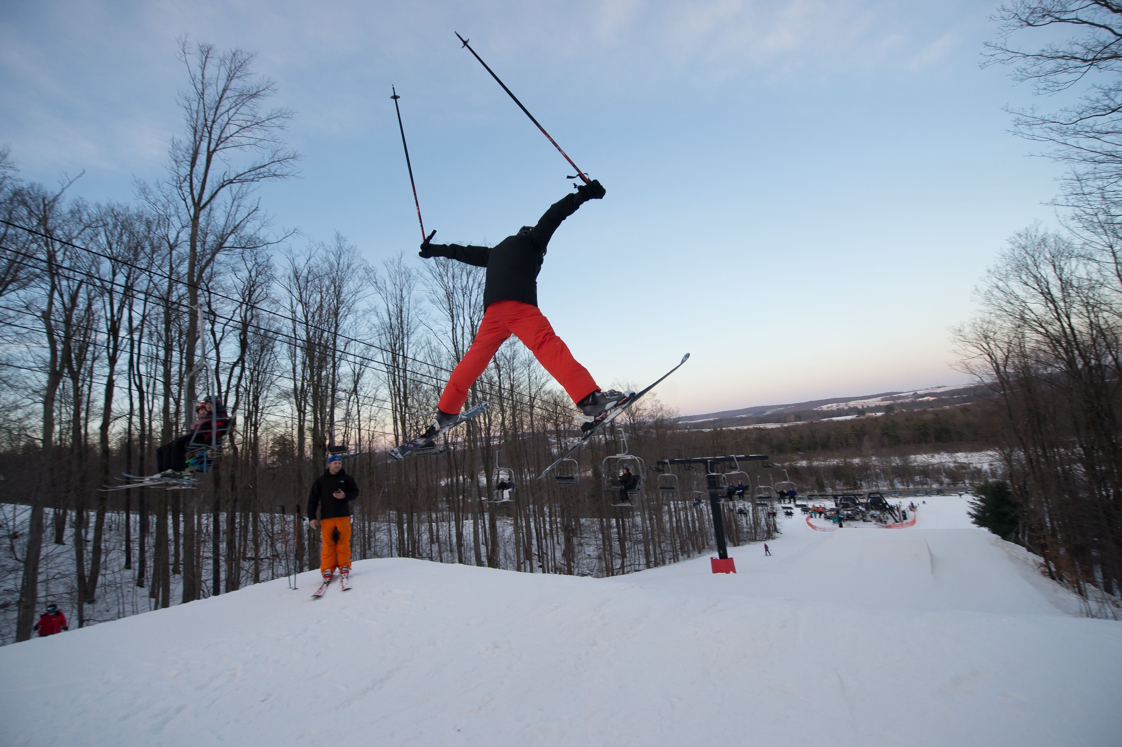 A ski slope with a person in mid-air performing a jump, surrounded by trees and ski lifts.