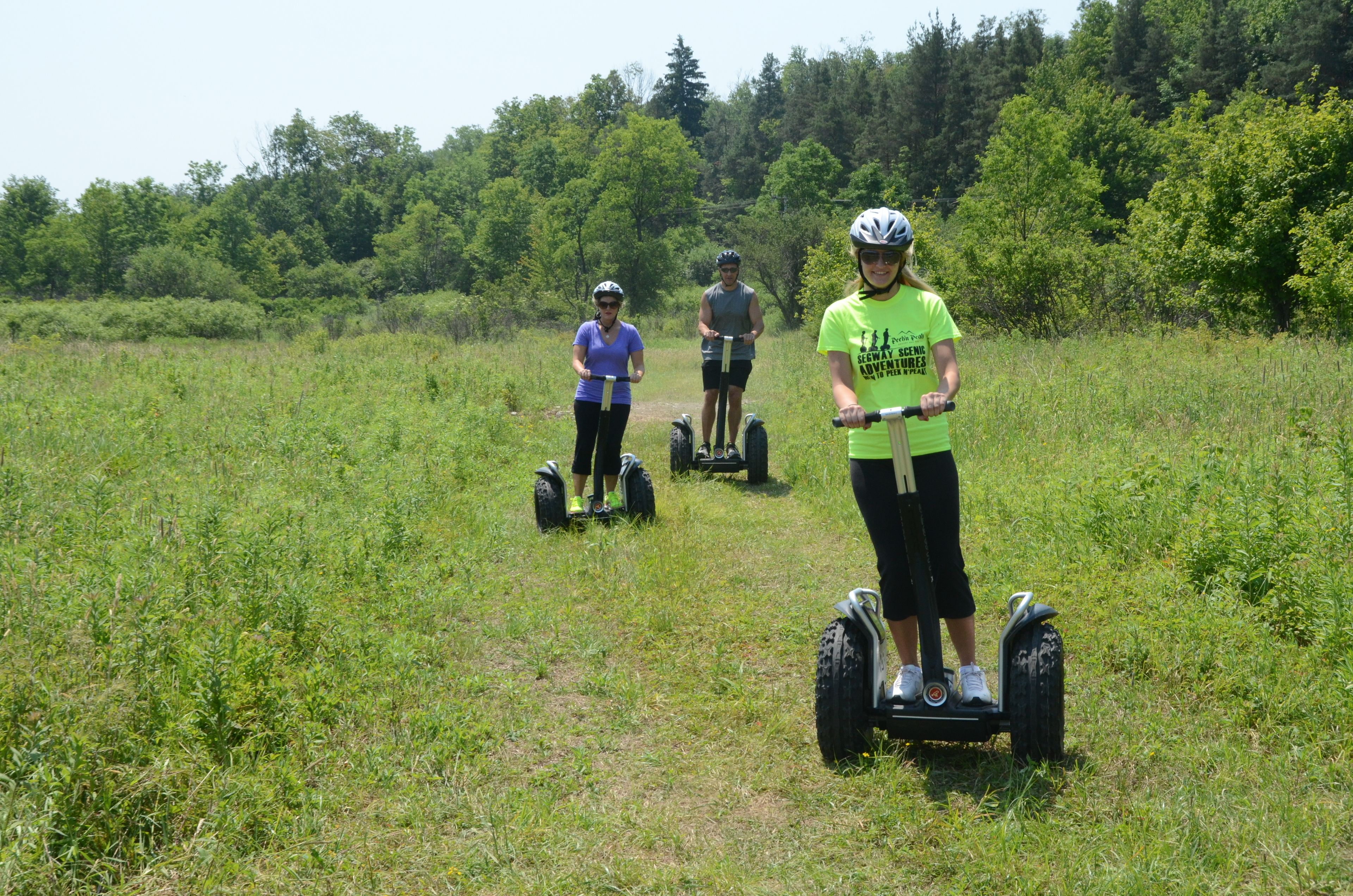 Three people on Segway-like devices in a grassy field with trees in the background.