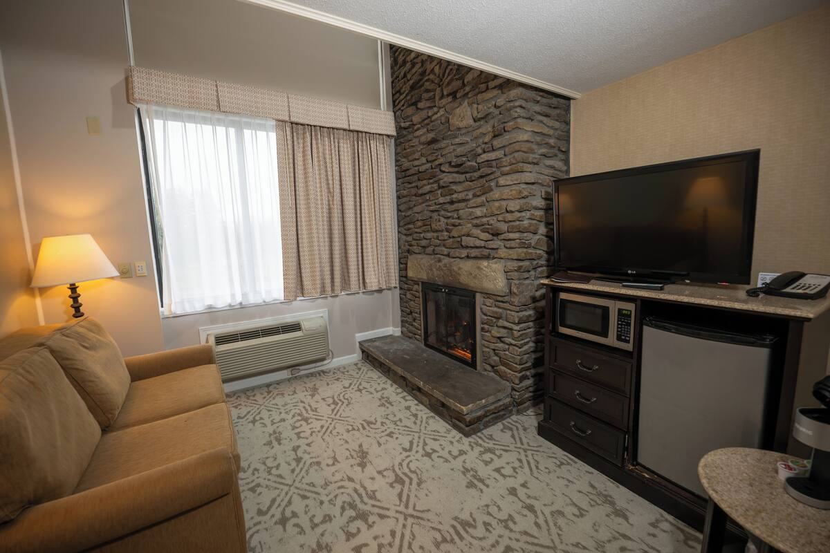 A living room with a stone fireplace, a flat-screen TV, a sofa, and a kitchen area with a microwave and refrigerator.