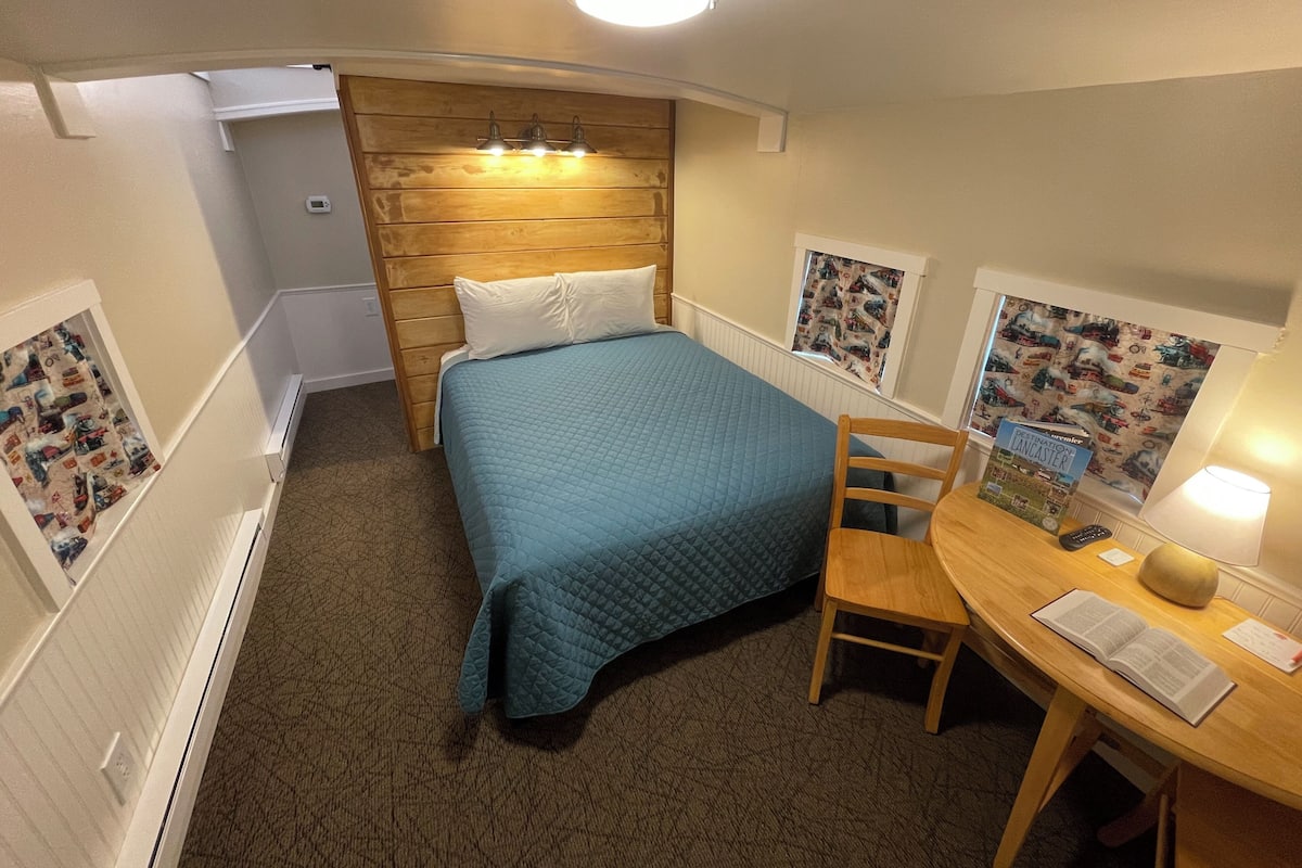 A compact room with a bed, a desk, and a chair. There is a window with a view of a building.