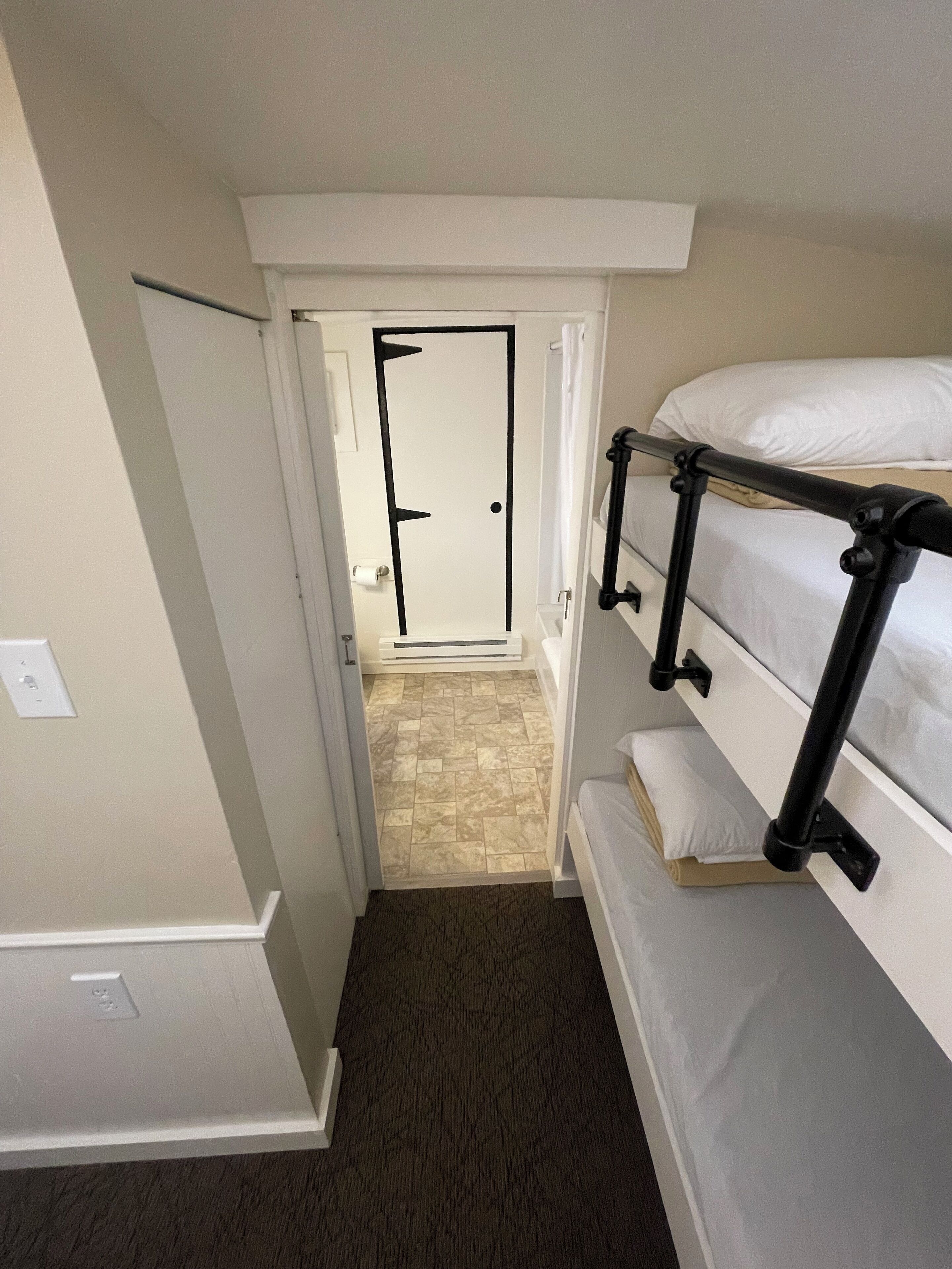 A narrow hallway leading to a door with a white frame and black accents, a bunk bed with a black metal frame, and a patterned carpet.