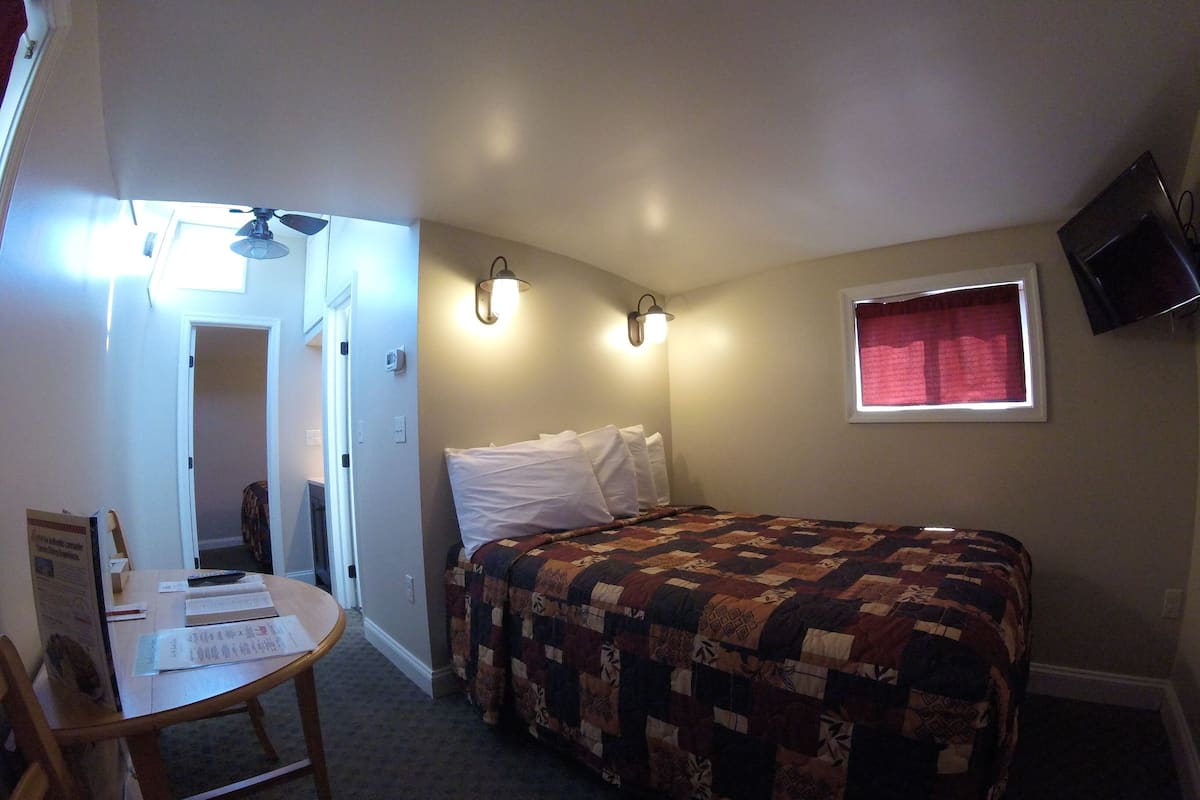 A bedroom with a bed, a desk, and a chair. There is a window with a red curtain, a wall-mounted TV, and a ceiling fan.