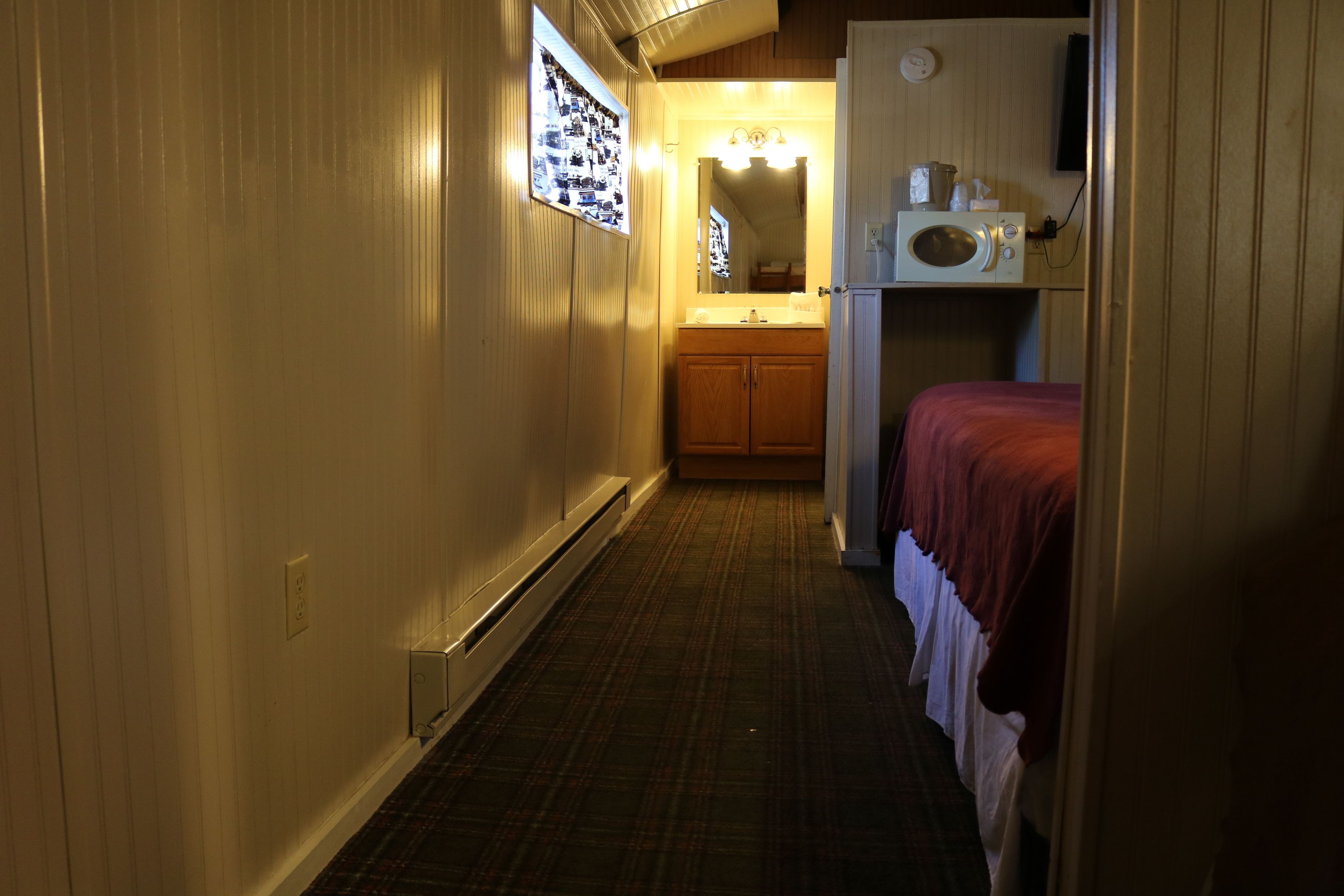 A narrow hallway with a wooden door on the right, a small bed with a red blanket, a microwave, and a sink with wooden cabinets.