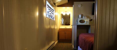 A narrow hallway with a wooden door on the right, a small bed with a red blanket, a microwave, and a sink with wooden cabinets.