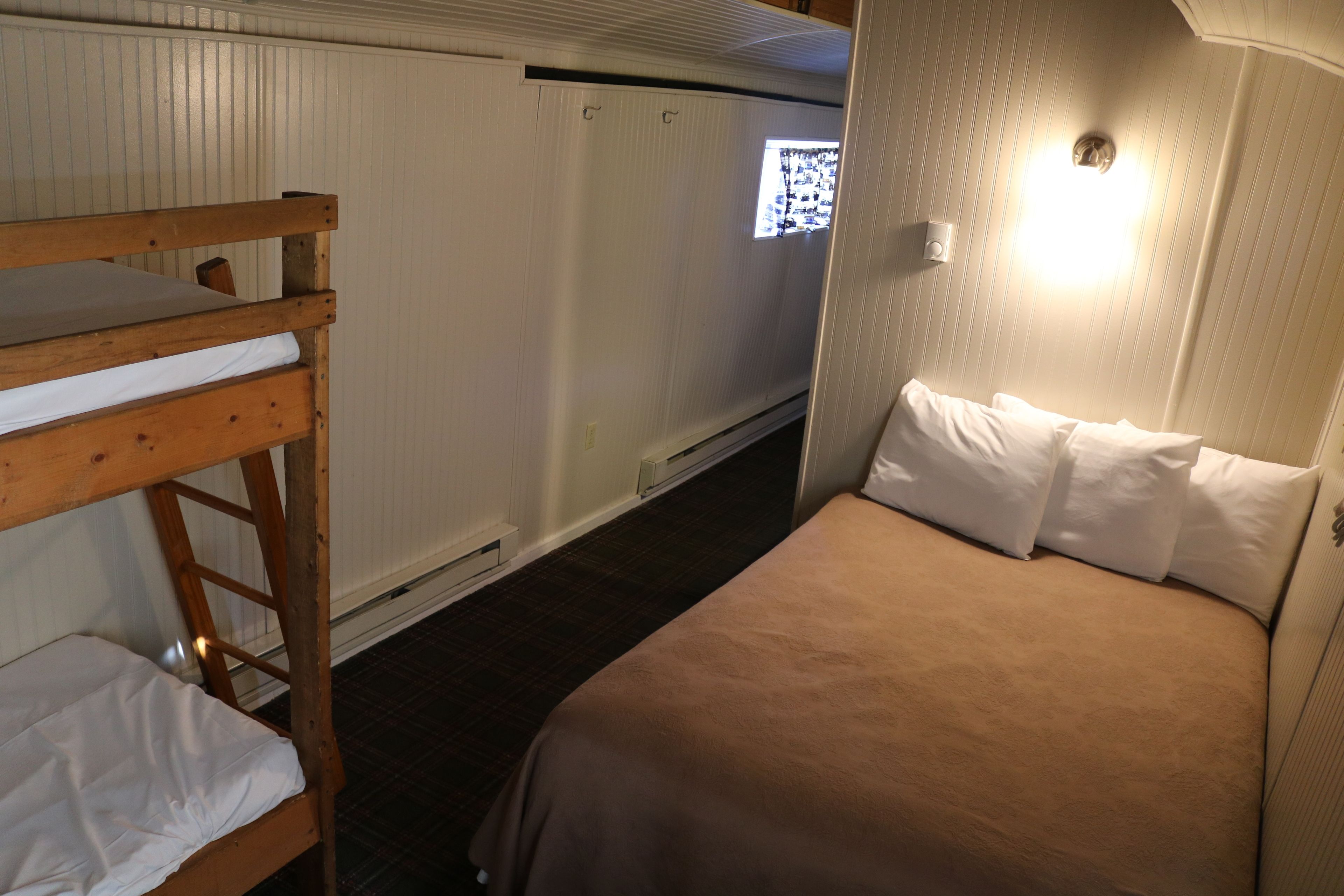 Una cama litera con una cama inferior y una cama individual con cabecera, ambas con ropa de cama blanca. Hay una ventana con persianas entreabiertas, dejando entrar luz natural.