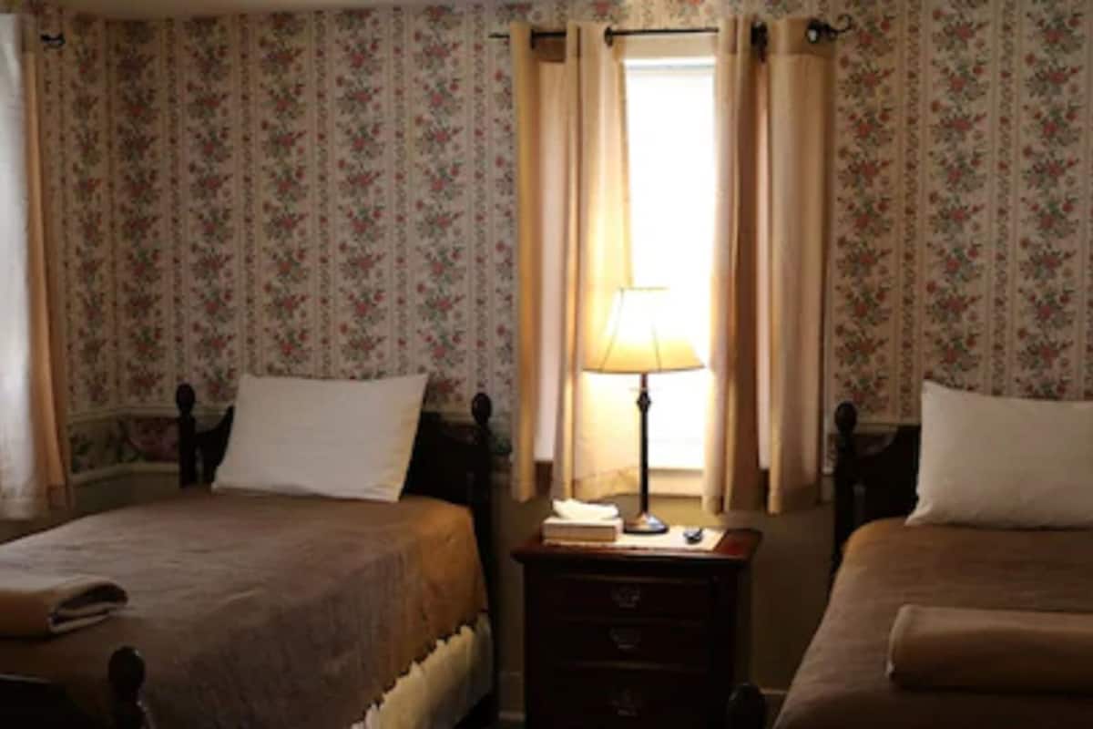 A room with two beds, floral wallpaper, a nightstand with a lamp, and a window with curtains.