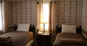 A room with two beds, floral wallpaper, a nightstand with a lamp, and a window with curtains.