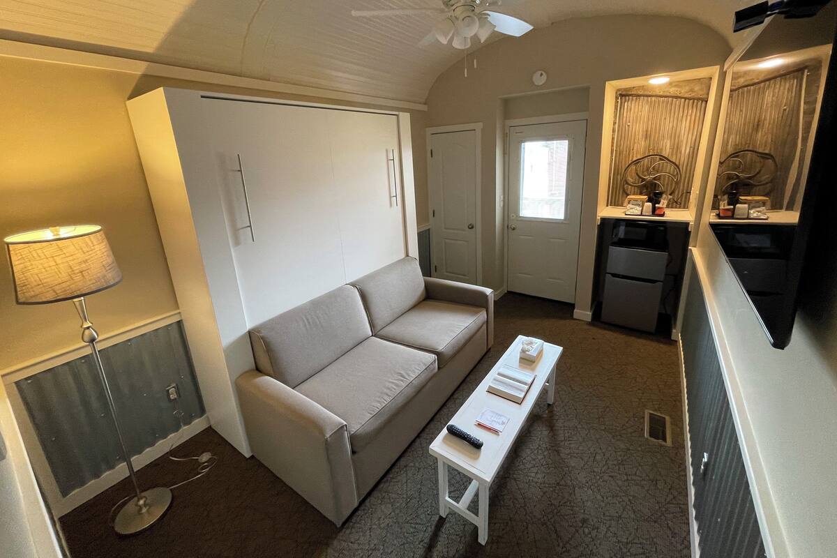 A compact living space with a sofa, a small table, a lamp, a ceiling fan, a kitchenette, and a door leading to another room.