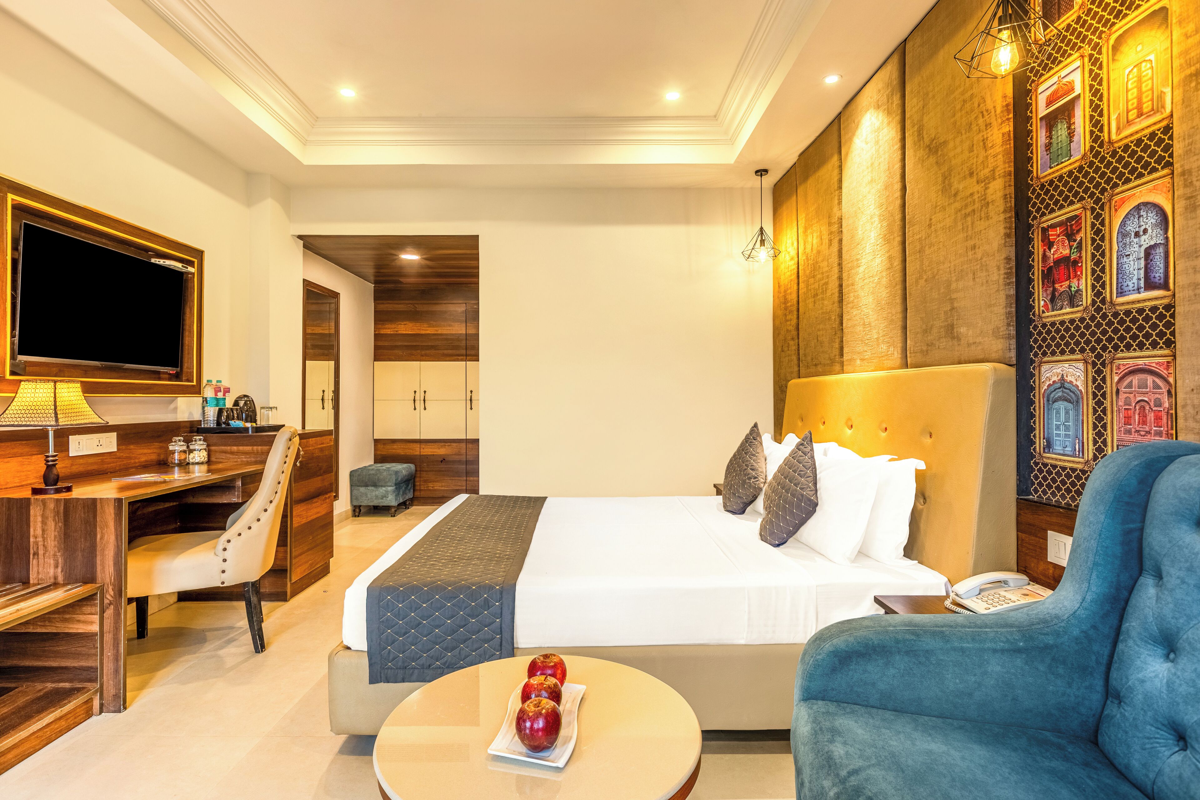 A hotel room with a bed, a desk, a chair, a table with apples, and a TV.